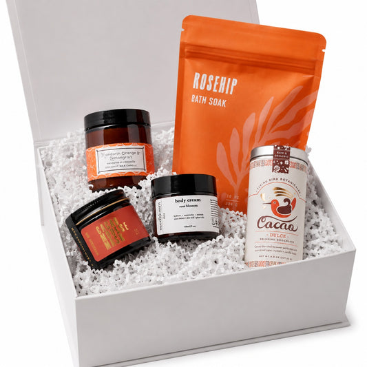 Box set of self-care products to create a blissful bath experience with the Bliss box including a Cacao mask, mandarin orange lemongrass candle, rose blossom hand lotion, cacao dulce for drinking, and the rosehip bath soak from URB Apothecary in the Here I Am gift box with white crinkle paper.