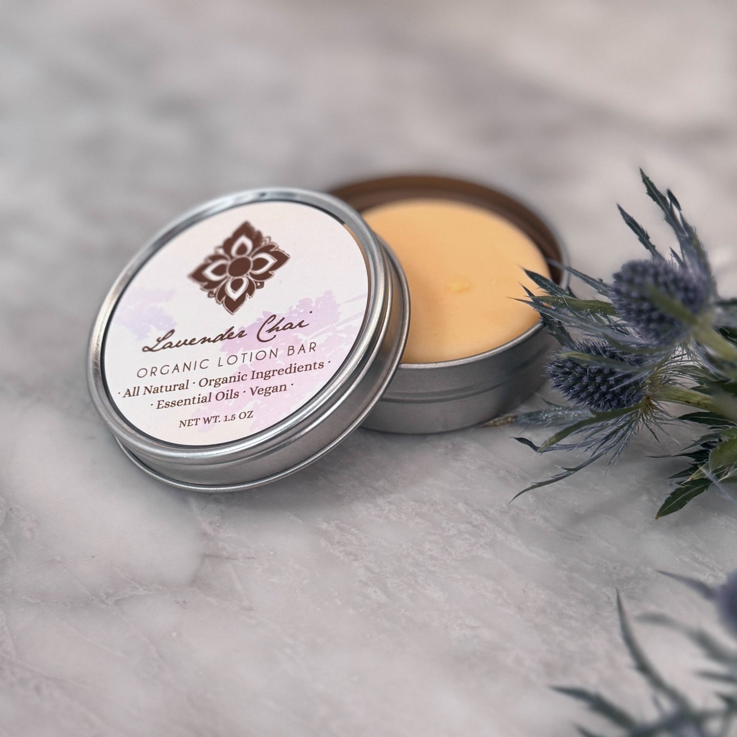 Open tin of Lavender Chai lotion bar on a counter next to fresh thistle