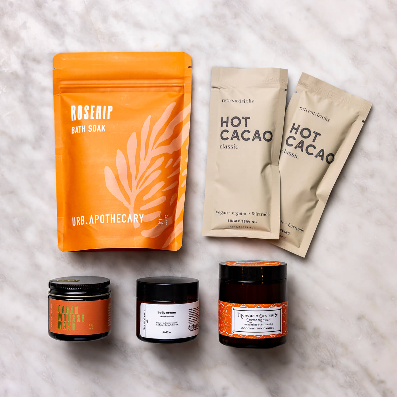 Collection of self-care products in the Blissful Bath set with the Cacao mask, hot cacao packet, mandarin orange lemongrass candle, floral hand lotion and rosehip bath soak.