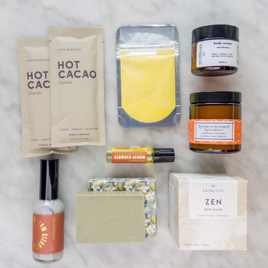 Collection of wellness products in the New Mom Self-Care Gift Box, including hot cacao, a turmeric face mask, and various other items on a light background.
