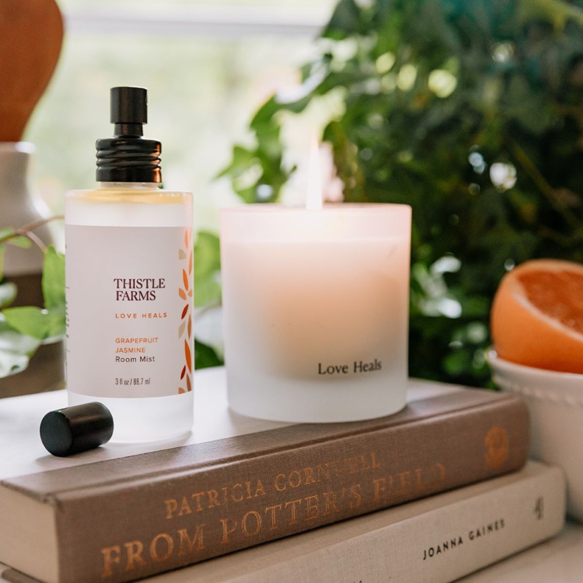 Thistle Farms aromatherapy room spray bottle and candle creating a zesty and uplifting atmosphere on books with plants in a sunny window