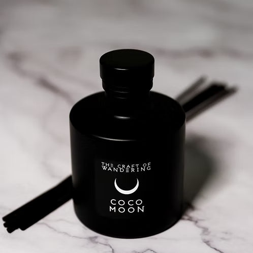 A matte black reed diffuser bottle with a matching black cap, labeled 'THE CRAFT OF WANDERING' and 'COCO MOON', set on a white marble surface with dark reeds visible behind it.