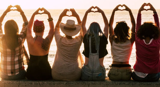 Line of women sitting on a wall facing the ocean with hands over heads in the "I love you" heart sign shape