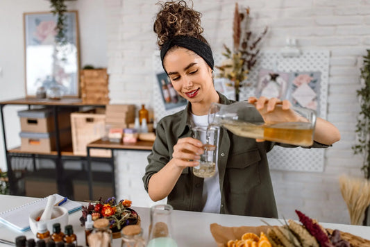 Woman pouring botanical oils from a pitcher into a beaker in a kitchen setting.