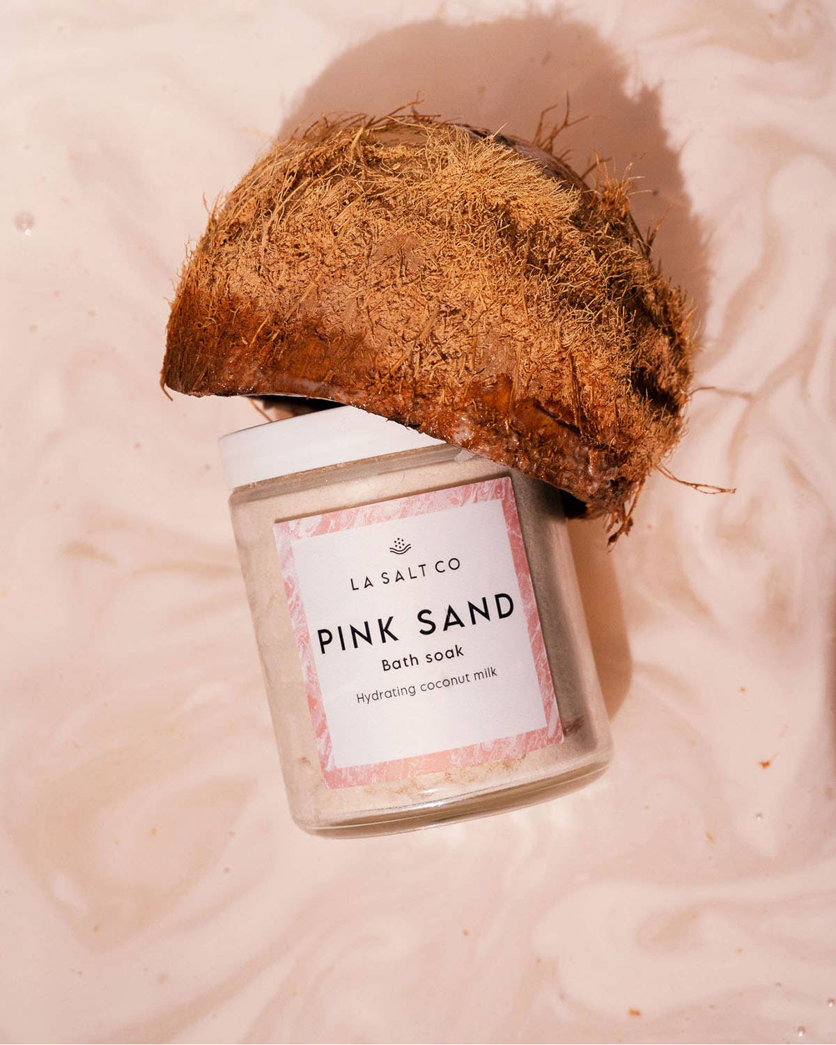 Jar of 'Pink Sand' coconut milk bath soak by La Salt Co with a coconut on a pink background