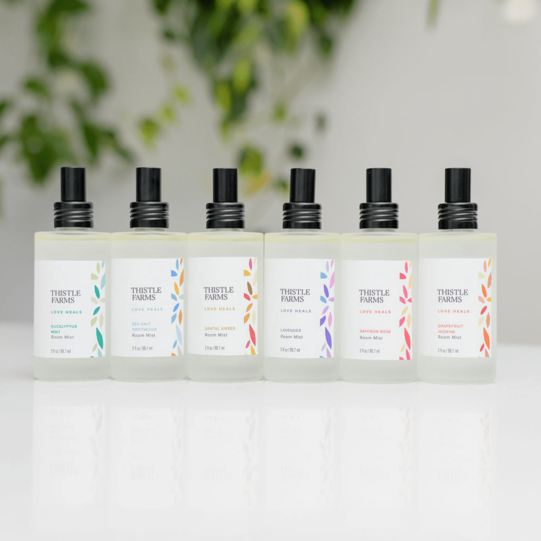 Set of Thistle Farms aromatherapy room spray bottles on a white surface with a blurred green plant background