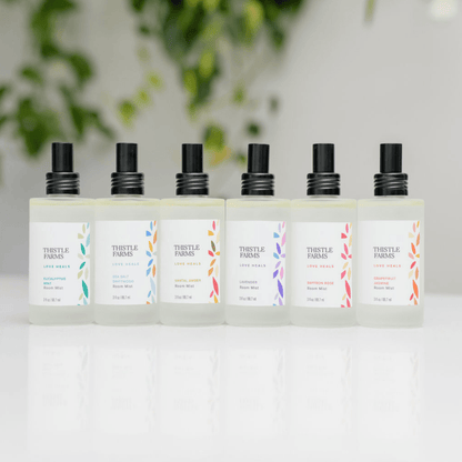 Set of Thistle Farms aromatherapy room spray bottles on a white surface with a blurred green plant background