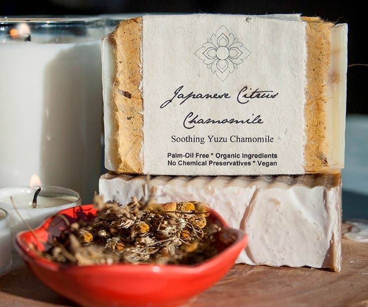 Bar of Citrus and Chamomile soap with a label on a wooden surface next to a bowl of dried herbs and a candle.