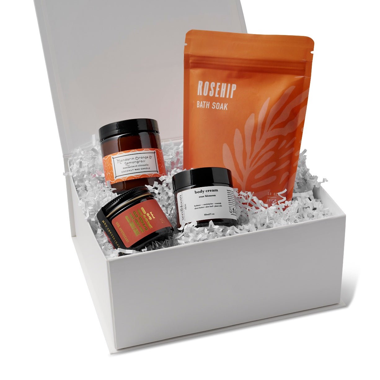 Box set of self-care products in the Bliss box with the Cacao mask, mandarin orange lemongrass candle, floral hand lotion and the rosehip bath soak from URB Apothecary in the Here I Am gift box with white crinkle paper. 