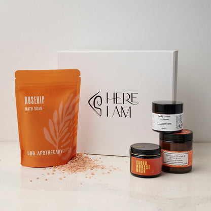 Box set of self-care products in the Bliss box with the Cacao mask, mandarin orange lemongrass candle, floral hand lotion and the rosehip bath soak from URB Apothecary with the Here I Am gift box in the background.