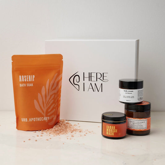 Box set of self-care products in the Bliss box with the Cacao mask, mandarin orange lemongrass candle, floral hand lotion and the rosehip bath soak from URB Apothecary with the Here I Am gift box in the background.