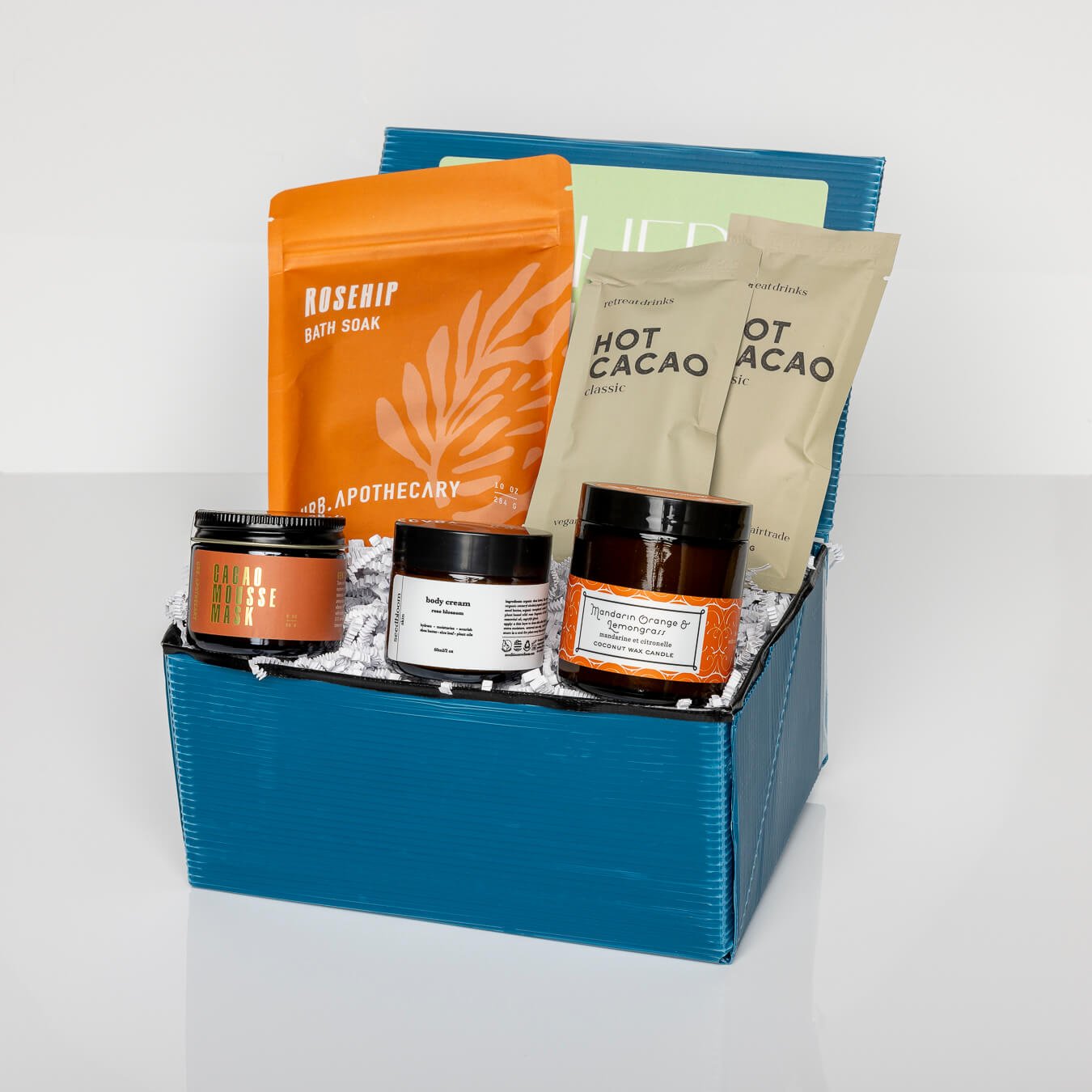 Blue box with cacao mask, hot cacao packet, orange lemongrass candle, hand lotion and rosehip bath soak.