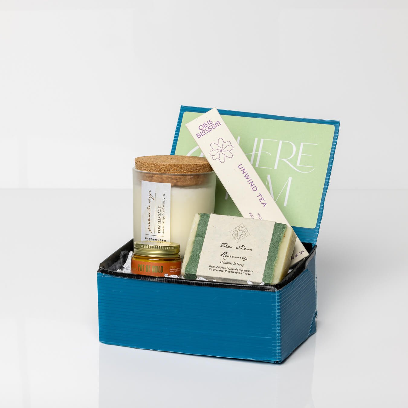 Blue gift box with a Pomelo Sage Candle, Pot of Gold Face Balm, Thai Lime Rosemary Soap, and Unwind Tea on white background.