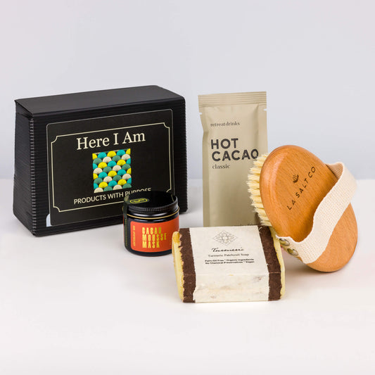 Cacao Cleanse Box from Here I Am, featuring a cacao mousse mask, hot cacao beverage, turmeric soap, and a body brush next to the iconic black box