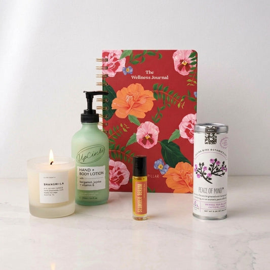 A curated wellness gift set on a white marble surface against a white wall, featuring a lit candle, bergamot green hand and body lotion, a small amber aromatherapy bottle, a silver tin of herbal tea, and a red floral spiral notebook titled 'The Wellness Journal'.