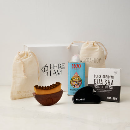 The products in the Flow Facial gift box including a tin of Blue Tansy and Apricot facial oil, a stone gua sha for face with a bag and box, and a gua sha brush with a drawstring bag with the premium Here I Am gift box in the background. 