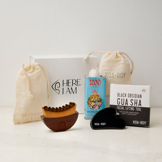 The products in the Flow Facial gift box including a tin of Blue Tansy and Apricot facial oil, a stone gua sha for face with a bag and box, and a gua sha brush with a drawstring bag with the premium Here I Am gift box in the background. 
