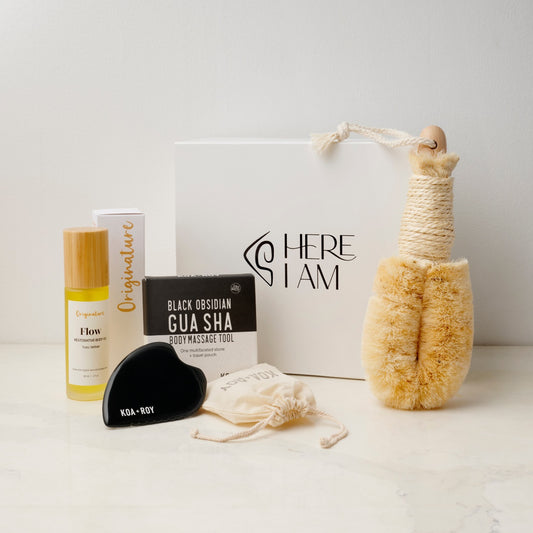 Collection of skincare and wellness products including a gua sha tool, body brush, and oil on a neutral background with the Here I Am Gift box. 