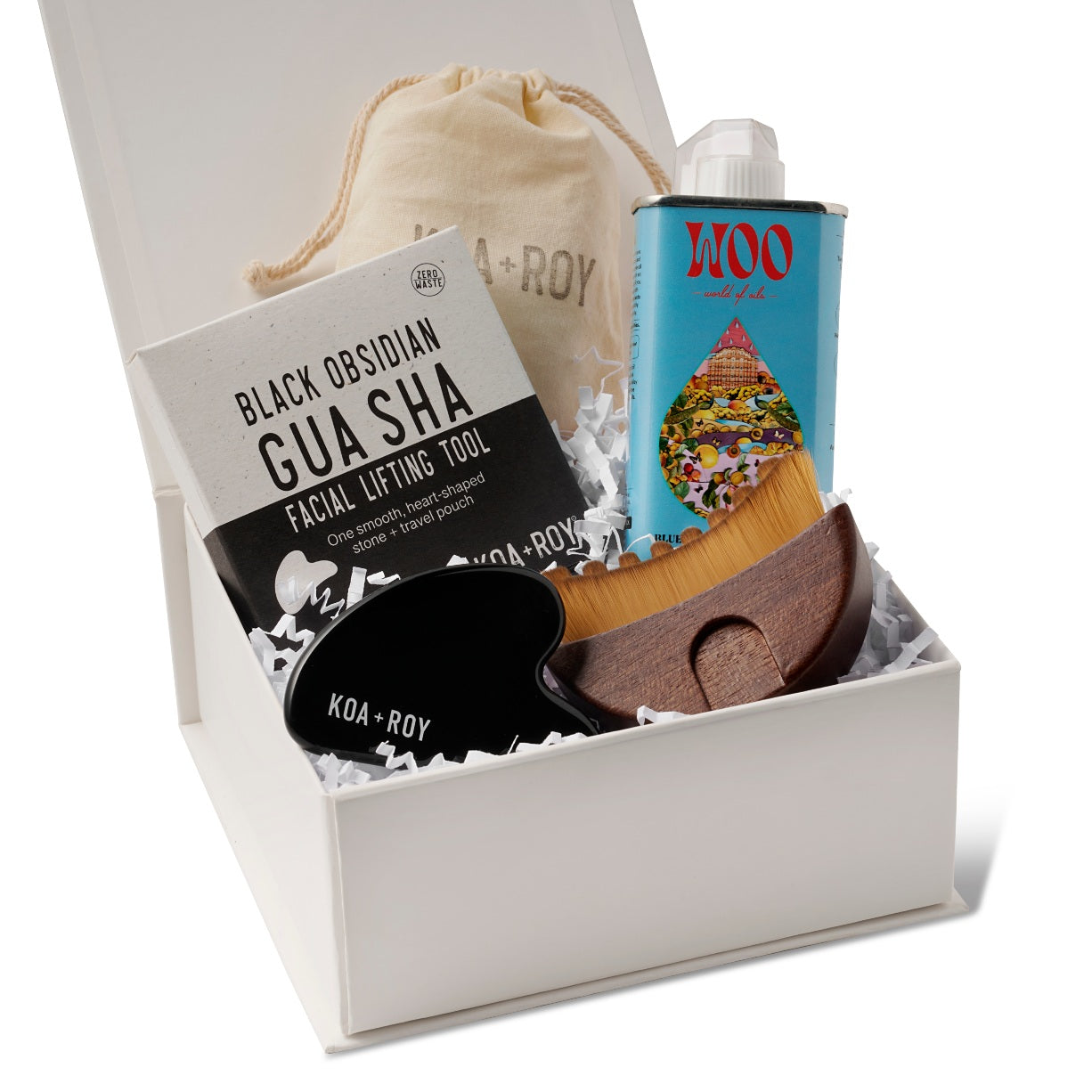 The products in the Flow Facial gift box including a tin of Blue Tansy and Apricot facial oil, a stone gua sha for face with a bag and box, and a gua sha brush with a drawstring bag in the premium Here I Am gift box with white crinkle paper. 
