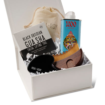 The products in the Flow Facial gift box including a tin of Blue Tansy and Apricot facial oil, a stone gua sha for face with a bag and box, and a gua sha brush with a drawstring bag in the premium Here I Am gift box with white crinkle paper. 