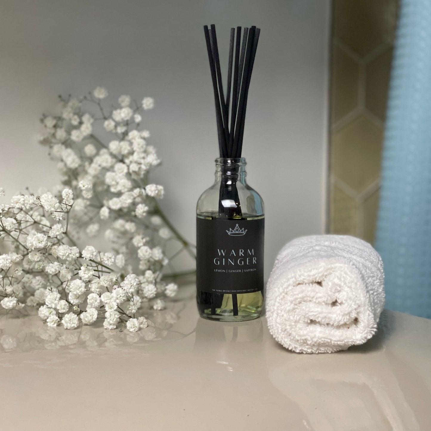 A diffuser bottle of Warm Ginger on a sink counter with white flowers and a washcloth