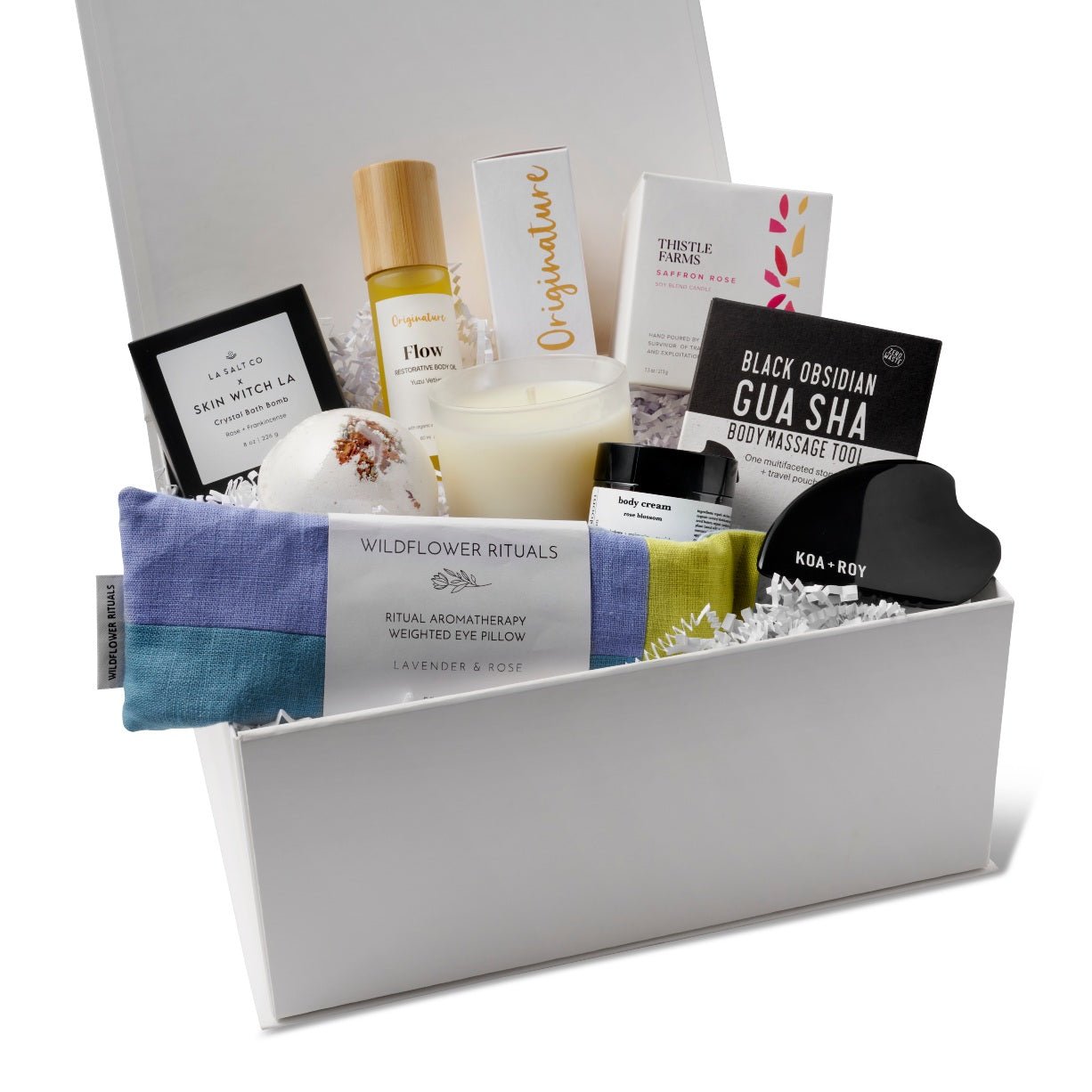A curated wellness gift set arranged inside a white gift box from HERE I AM, with a blue and yellow eye pillow, an essential oil candle, a gua sha body massage tool, a bath bomb, a small jar of lotion, and a bottle of skin oil.