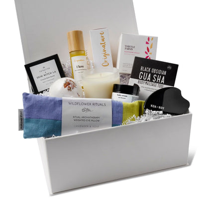 A curated wellness gift set arranged inside a white gift box from HERE I AM, with a blue and yellow eye pillow, an essential oil candle, a gua sha body massage tool, a bath bomb, a small jar of lotion, and a bottle of skin oil.