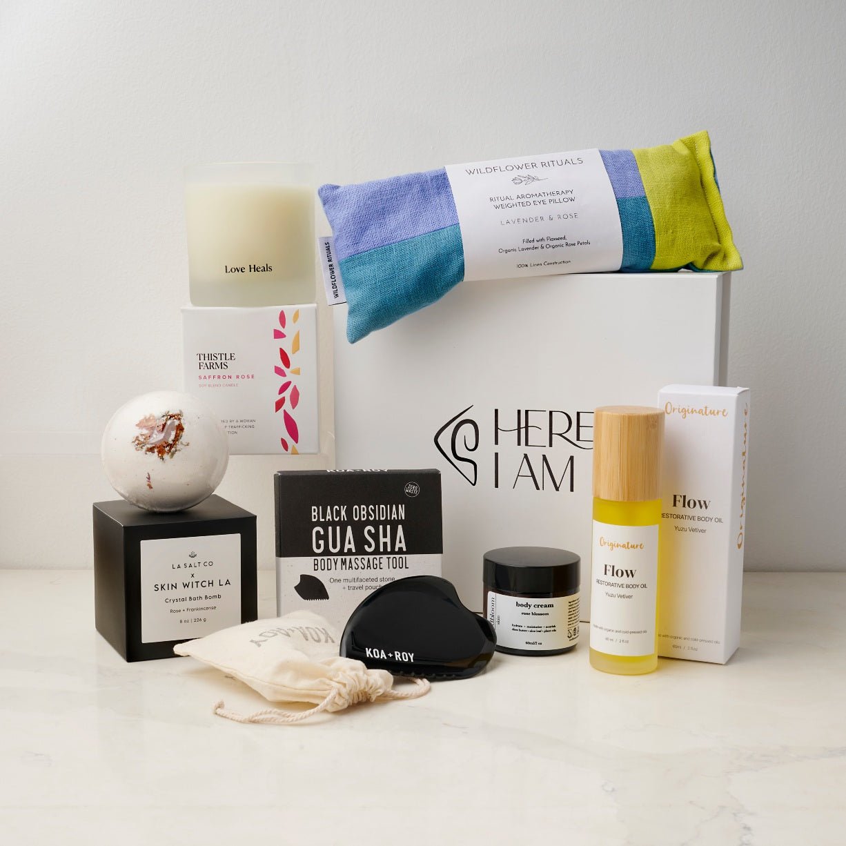 A curated wellness gift set arranged on a light marble surface against a white wall. The set includes a white box labeled HERE I AM, a blue and yellow eye pillow, an essential oil candle, a gua sha body massage tool, a bath bomb, a small jar of lotion, and a bottle of skin oil.