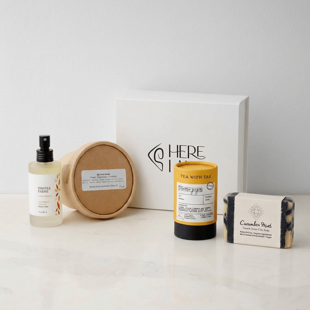 Collection of skincare and personal care products including essential oil room spray, foot soak, soap, and tea with a Here I Am gift box on a white background.