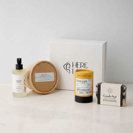 Collection of skincare and personal care products including essential oil room spray, foot soak, soap, and tea with a Here I Am gift box on a white background.