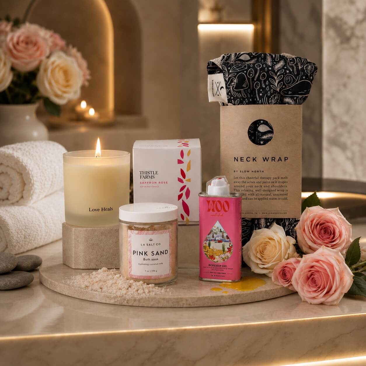 Gifting set with candles, bath salts, rosehip oil, and a neck wrap on a marble surface with flowers.