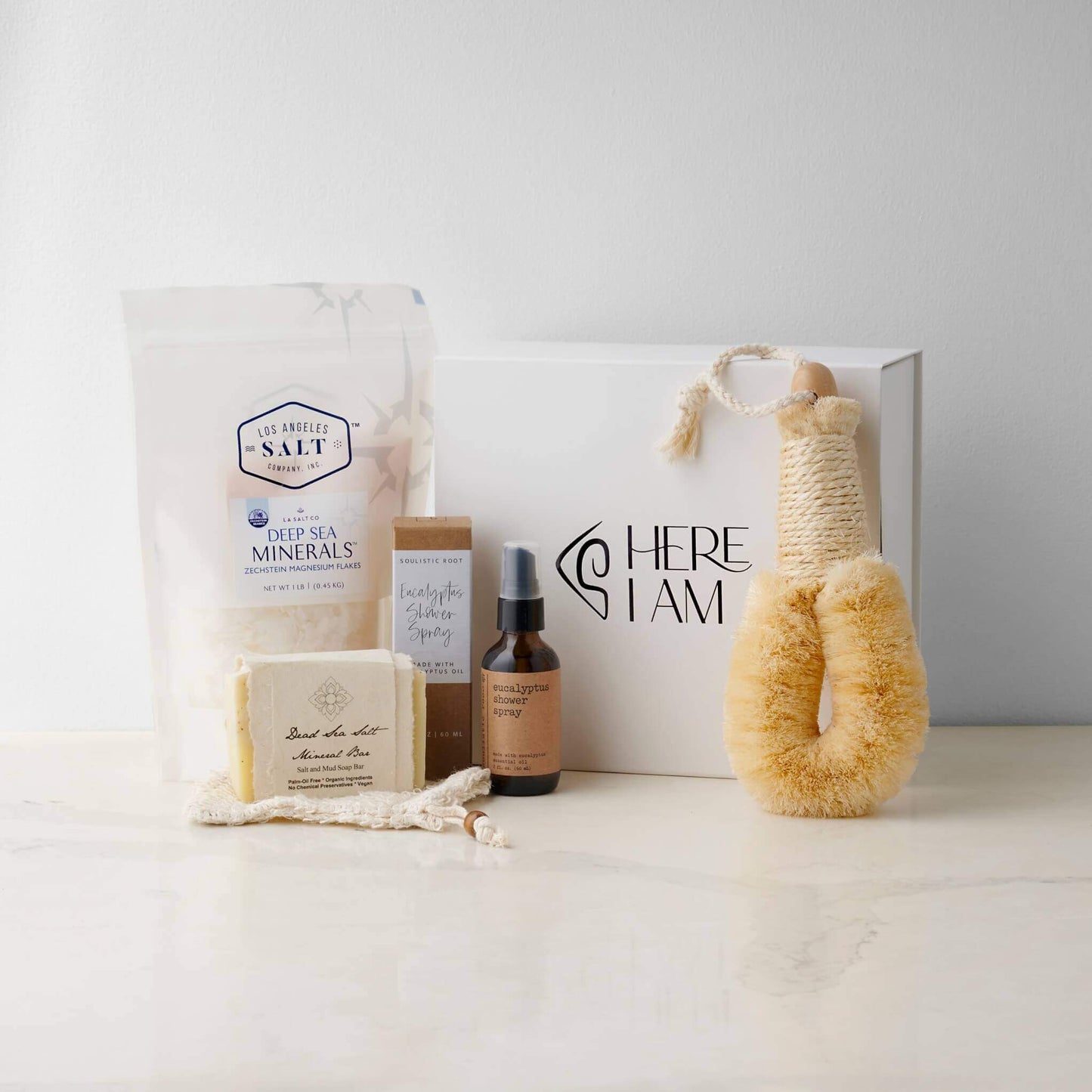 Contents in the Magnesium Bath Box, including Zechstein sea salts, body brush, shower spray, and soap with a Here I Am giftbox in the background.