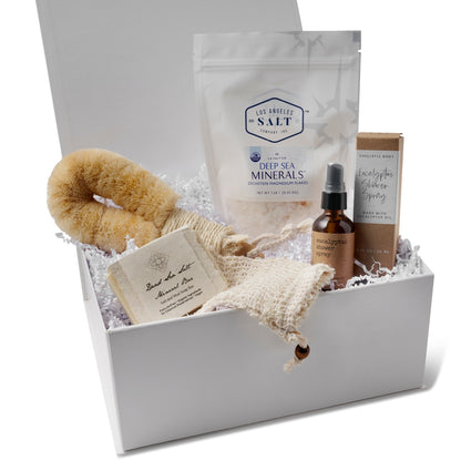 Contents in the Mineral Rescue Bath Box, including Zechstein sea salts, dry body brush, shower spray, and soap in a Here I Am gift box with white crinkle paper.