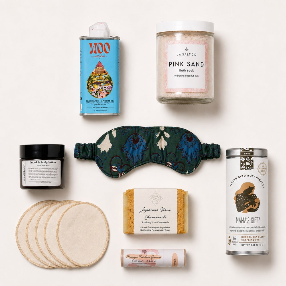 A curated flat lay of wellness and beauty products on a clean white background, including a blue tin of WOO blue tansy oil,  a jar of Pink Sand bath soak, a dark green floral sleep mask, a jar of rose blossom body lotion, a bar of Japanese Chamomile soap, a tube of lip balm, and a stack of round reusable face pads.