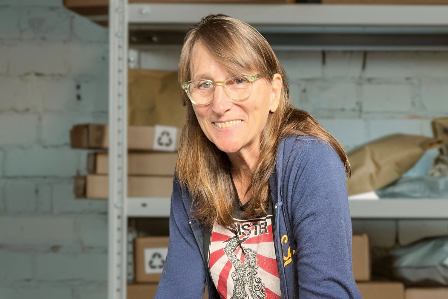Renee stands behind a table in her warehouse with cardboard boxes on metal shelving in the background.