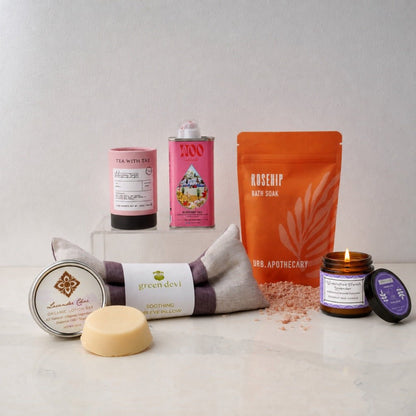 A curated spa gift set arranged on a light marble surface against a neutral gray background. The set includes a Green Devi soothing eye pillow, a lavender lotion bar, a rosehip bath soak pouch, a hibiscus rose tea, a pink tin with rosehip face oil, and a small lavender essential oil candle with a purple label.