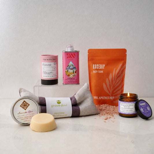 A curated spa gift set arranged on a light marble surface against a neutral gray background. The set includes a Green Devi soothing eye pillow, a lavender lotion bar, a rosehip bath soak pouch, a hibiscus rose tea, a pink tin with rosehip face oil, and a small lavender essential oil candle with a purple label.