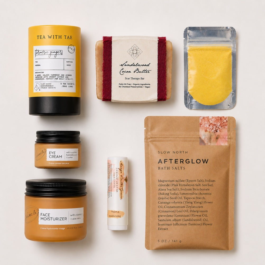 A curated flat-lay of six skincare and beauty products from the Skin Renewal Self-Care Gift Box on a clean white background, including jars of face cream and moisturizer, a bar of soap, a tube of lip balm, a pouch of bath salts, and a cardboard tube of turmeric ginger tea.
