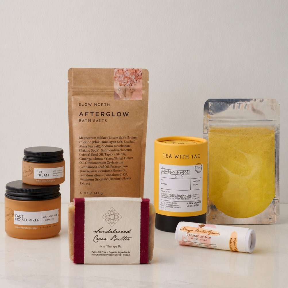 A curated wellness products from the Skin Renewal Self-Care Gift Box featuring Slow North bath salts, a tea with tae turmeric ginger tea, a handcrafted sandalwood cocoa butter bar, and moisturizers, arranged on a white surface against a neutral off-white background.