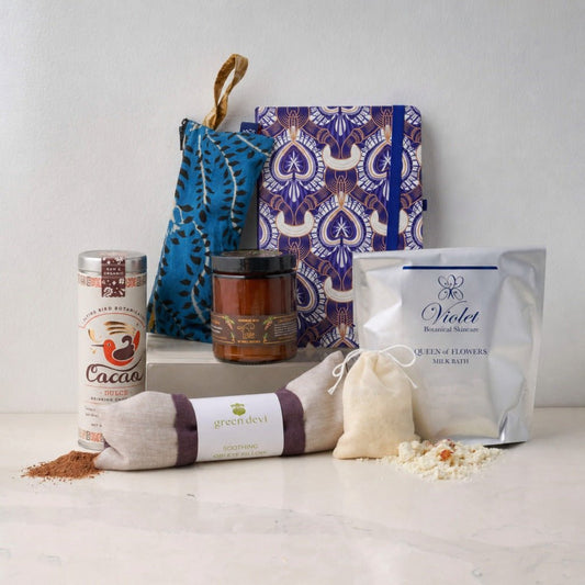 A curated gift set arranged on a light stone surface against a white wall, including a silver canister of cacao drinking chocolate, a patterned blue and purple notebook, a glass apothecary jar candle, a silver bag with goat's milk bath sachets, a linen eye pillow with a purple band, and scattered powder.