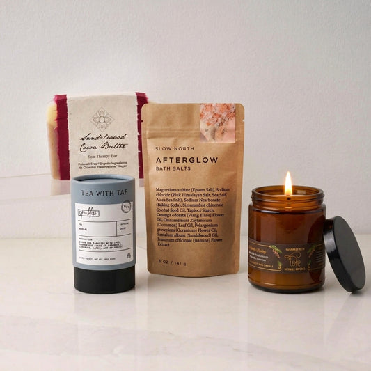 Collection of wellness products from the Zen Moment box, including a handcrafted cocoa butter soap bar in a red and white wrapper, a brown pouch of Bath Salts, a tube of herbal tea, and an amber glass candle jar on a white marble counter.