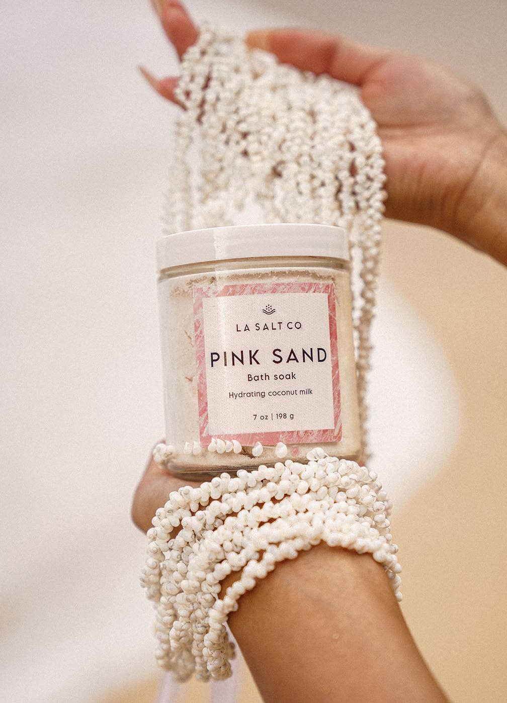 Person holding a 'Pink Sand' coconut milk bath soak with decorative beads against a neutral background