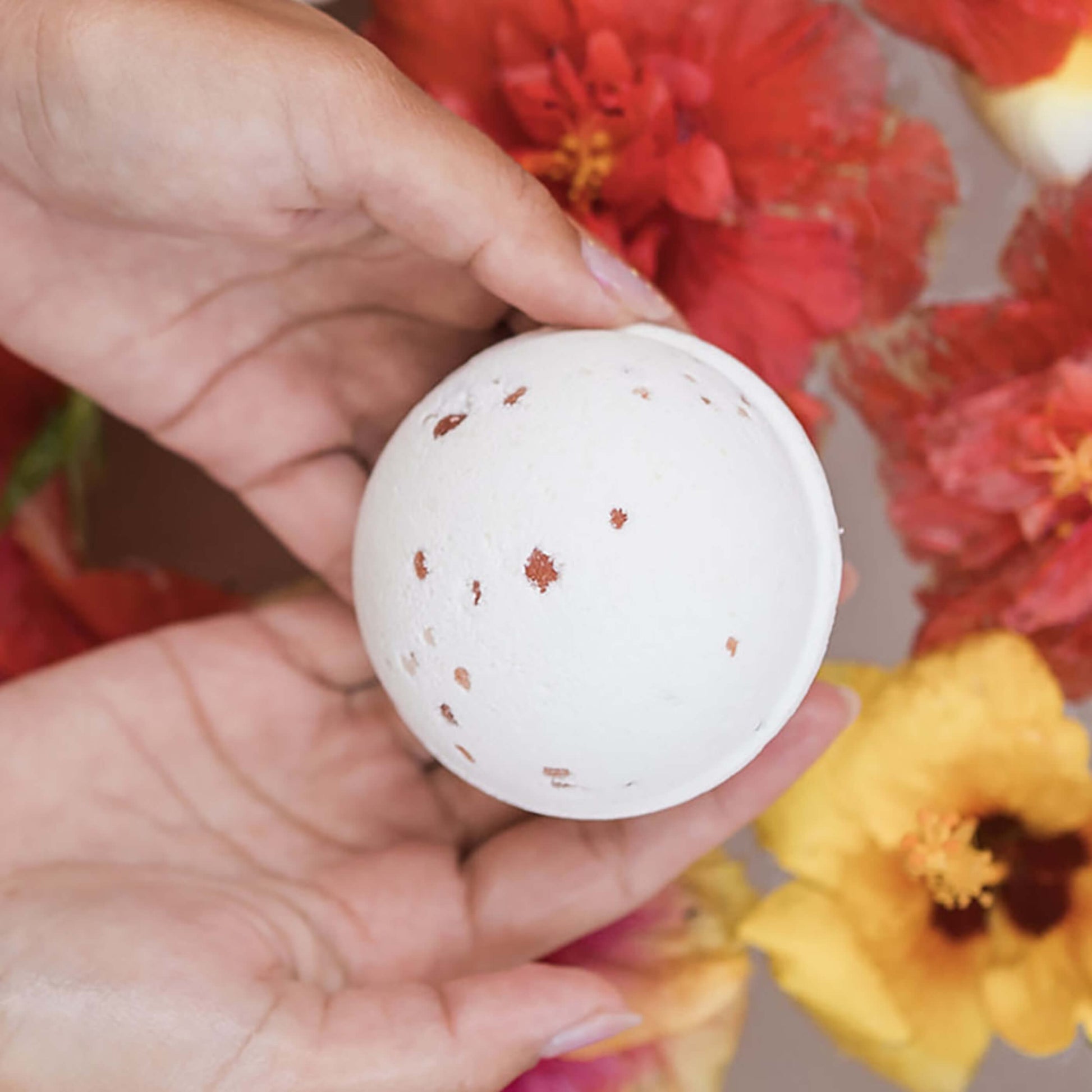 Two hands holding the Zen Bath Bomb over a tub of water with colorful hibiscus flowers
