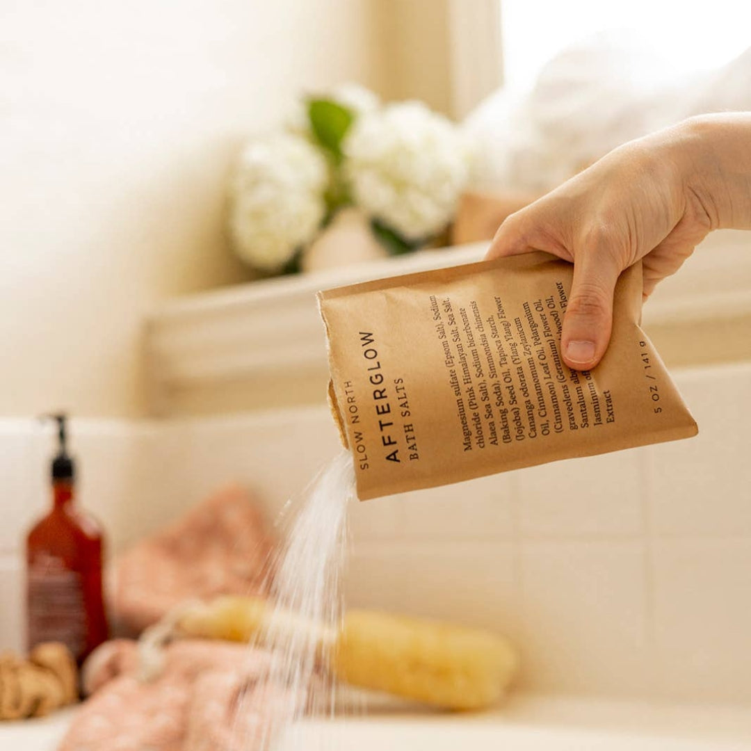 A hand holding a brown paper packet of Slow Nortj AfterGlow Bath Salts, pouring the salts into a bathtub with a sisal brush and pink towels nearby.