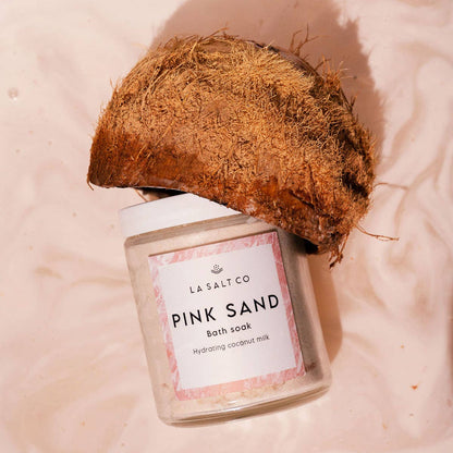 Jar of 'Pink Sand' coconut milk bath soak by La Salt Co with a coconut on a pink background