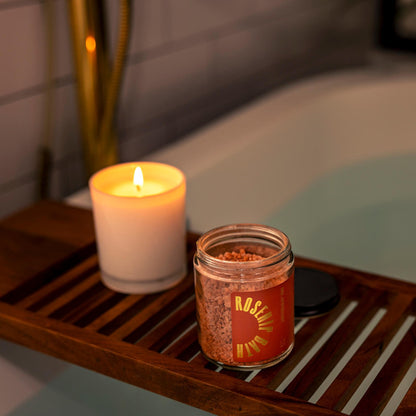 A serene setting with an open jar of the Rosehip Bath Salts on a bamboo shelf over a full bath next to a burning candle