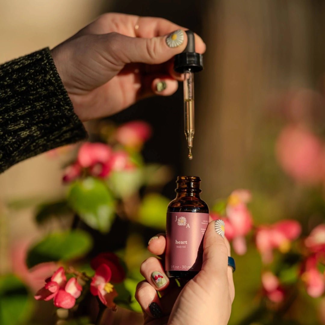 A close-up lifestyle shot of a person holding an amber glass dropper bottle and dispensing a drop of floral body oil. The background is a soft, warm-focus outdoor scene with pink flowers and green leaves.