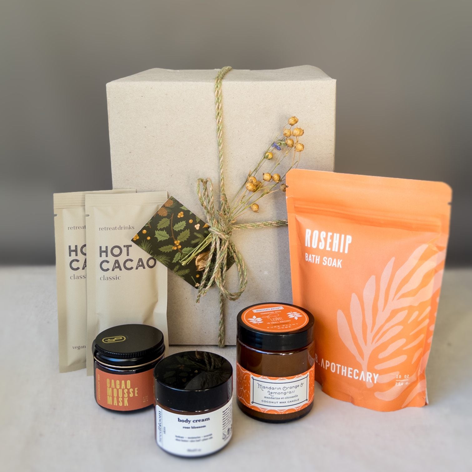 Blissful Bath Gift Box with the Cacao mask, hot cacao packet, mandarin orange lemongrass candle, floral hand lotion and rosehip bath soak in front of a wrapped gift box. 