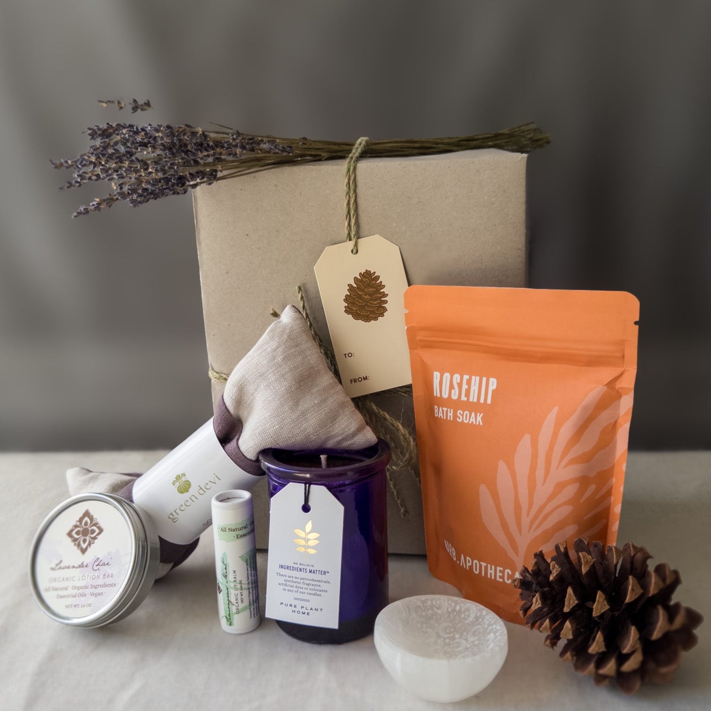 The Box contents for the Empty Nester Survival Kit with eye pillow, bath salts, and candle in front of a wrapped gift box with lavender and pinecone.