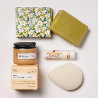 Skincare products in the Fresh Face box including lip balm, a face moisturizer, soap made with sandalwood essential oil, and a konjac sponge on a white tile surface.